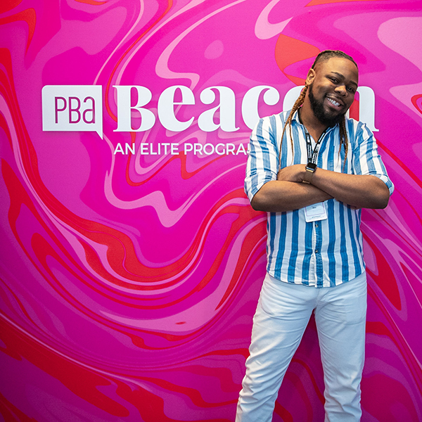 Man posing in front of a pink Beacon branded sign with a blue stripped shirt