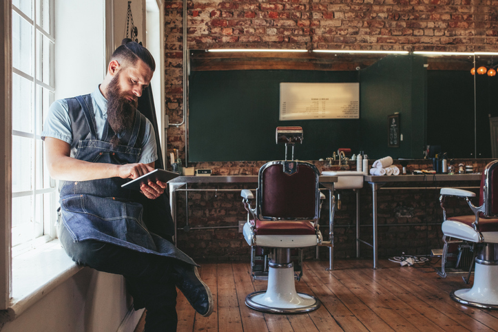 Barber with digital tablet sitting on window sill of salon. Hairdresser organizing his business using latest technology.