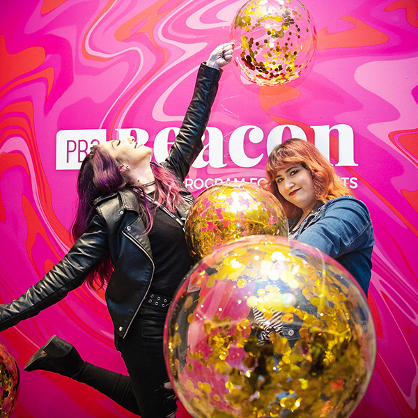 Two women holding balloons with gold glitter in them in front of pink banner