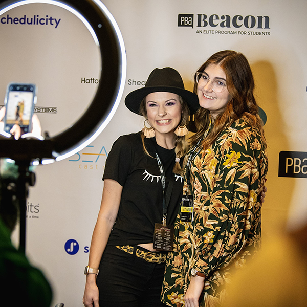 Two women posing in front of a branded Beacon banner ring light and cameras